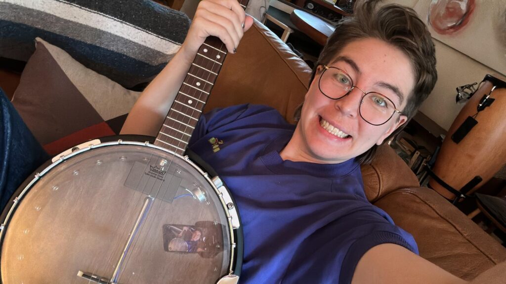 Paterson- a white, non-binary, 30 year old- is smiling excitedly while holding a banjo.